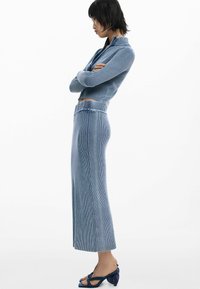 Vrouw in een blauwe geribde gebreide zip-up sweater en een bijpassende lange rok, staand in profiel met de armen gekruist en navy hakken aan.