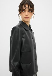 Schwarze Kunstlederjacke mit Kragen, Vorderknöpfen und einer aufgesetzten Tasche. Glatte Textur und strukturiertes Design.