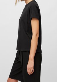Persona vestida con una camiseta de manga corta negra lisa y pantalones cortos negros hasta la rodilla, fotografiada de lado contra un fondo liso.