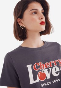 T-shirt en coton gris avec le texte « Cherry Love », présentée avec un graphique de pomme rouge. Des boucles d'oreilles dorées en forme de cœur complètent le look du modèle.