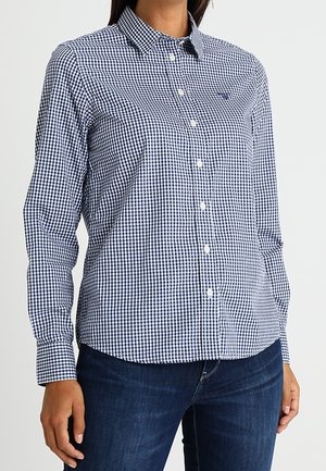 Kobieta w dopasowanej, długorękawowej koszuli w niebiesko-białą kratę oraz ciemnoniebieskich dżinsach, stojąca na tle jednolitego tła.