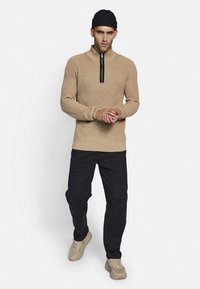 Beżowy sweter wykonany na drutach z wysokim kołnierzem i czarnym suwakiem, w zestawieniu z czarnymi spodniami i jasnobeżowymi trampkami. Model stoi na białym tle.