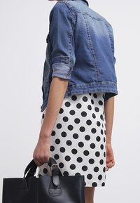 Veste en denim au design court, manches retroussées ; robe blanche à gros pois noirs ; tenant un sac à main en cuir noir.