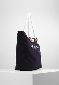 Marinblåt tote väska med vita rephandtag, prydd med en logotyp tryckt i vitt. Tillverkad av canvas med en slät textur och en rektangulär form.