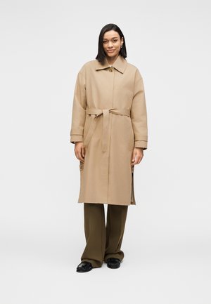 Beiger Trenchcoat mit Gürtel, Kragen, langen Ärmeln und seitlichen Knöpfen, kombiniert mit olivgrünen weiten Hosen und schwarzen Schuhen.