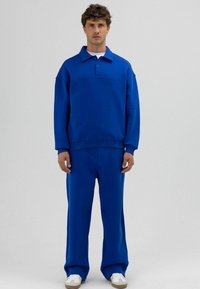 Blauw jersey sweatshirt met een polokraag en knopen aan de voorkant, gecombineerd met bijpassende blauwe broek met wijde pijpen. Soepele structuur en ontspannen pasvorm.