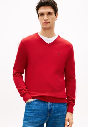 Uomo che indossa un maglione rosso con scollo a V sopra una T-shirt bianca e jeans blu, in piedi con una mano in tasca.