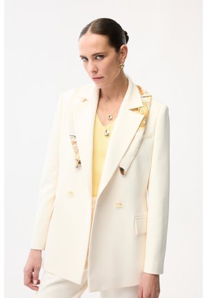 Kvinde iført en cremefarvet dobbeltradet blazer med blomsterdetaljer over en gul top, accessoriseret med guldbøjleøreringe og lagdelte halskæder.