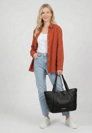 Junge Frau trägt ein rostfarbenes Hemd mit Knöpfen, weißes Top, helle Jeans, weiße Sneakers, hält eine große schwarze Tragetasche und lächelt.