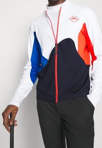 Zip-up sportjacka i vitt, blått, rött och marinblått. Har färgblock, hög krage och en framtida dragkedja med kontrasterande detaljer.