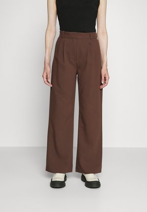 Osoba nosząca brązowe spodnie z wysokim stanem i szerokimi nogawkami z zakładkami, czarny bezrękawnik i białe buty z czarnymi podeszwami.