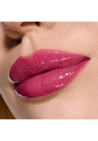 Labbra lucide color prugna con una texture liscia; una leggera brillantezza mette in risalto la pienezza, su uno sfondo di tonalità della pelle morbida e naturale.