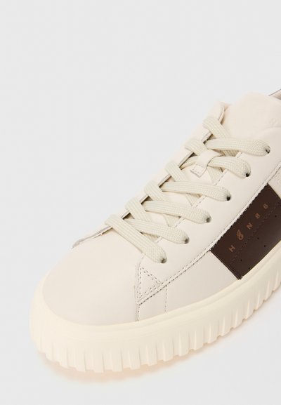 Primo piano di una sneaker crema con suola spessa, lacci crema e una striscia laterale marrone con un piccolo logo e il testo "HON86".