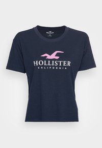 Mörkblå bomull t-shirt med rund halsringning, korta ärmar och en vit och rosa "HOLLISTER"-logotyp centrerad på framsidan.