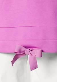 Top de algodón rosa con dobladillo redondeado y detalle de lazo en la cintura, combinado con una tela blanca, texturizada y lisa.