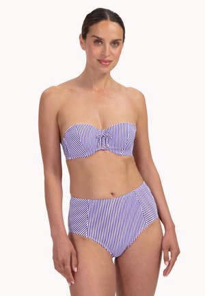 Frau trägt einen blau-weiß gestreiften trägerlosen Bikini mit vertikalen Streifen und einer Schleifendetail am Oberteil, steht vor einem einfarbigen hellen Hintergrund.