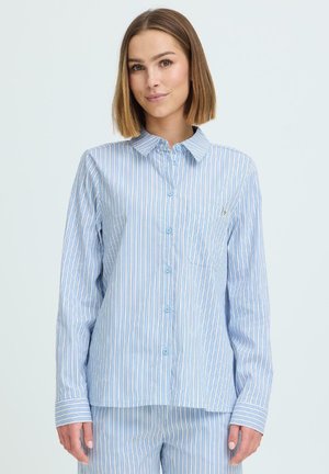 Mujer vestida con un conjunto de pijama de botones con rayas verticales en azul claro y blanco, de pie frente a un fondo claro y liso.