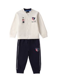Chaqueta blanca con cremallera con gráficos de fútbol y baloncesto, combinada con pantalones deportivos azul marino con rayas y acentos de logotipo similares.