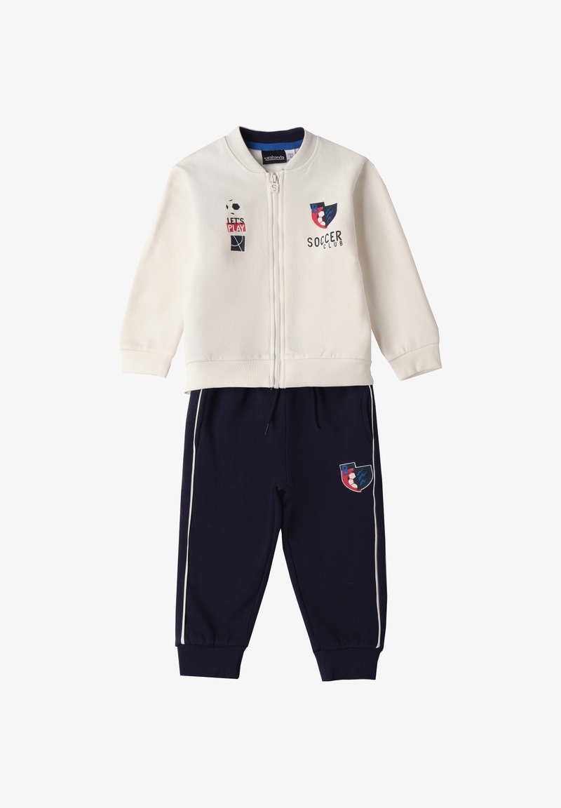 Veste zippée blanche avec des graphiques de football et de basket-ball, associée à un pantalon de jogging marine avec des rayures et des accents de logo similaires.