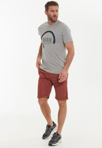 Grå t-shirt med svart grafisk design, matchad med röda shorts och grå sneakers. Modellen står med en avslappnad hållning och upprullade shorts.