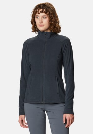 Femme aux cheveux bouclés portant une veste polaire sombre zippée et un pantalon gris, debout devant un fond clair uni.