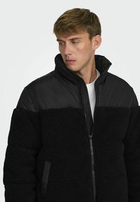 Chaqueta puffy negra con un panel inferior de forro polar texturizado, parte superior de nailon liso, cuello alto y cierre frontal con cremallera. Cuenta con bolsillos laterales.