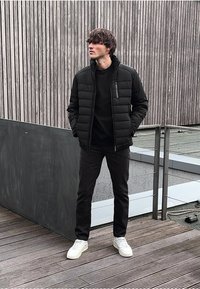 Černá péřová bunda s kapsou na zip, černé tričko, tmavé džíny a bílé tenisky. Stojí na dřevěných prknech s texturovaným pozadím.