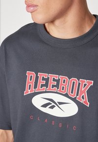 T-shirt en coton gris présentant un grand logo « REEBOK » imprimé en rouge et un ovale blanc en dessous, avec « CLASSIC » en texte plus petit. Manches courtes.