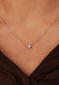 Collier en chaîne en argent avec un petit pendentif rond en pierre transparente, reposant sur un décolleté de teinte chair avec un haut marron visible.
