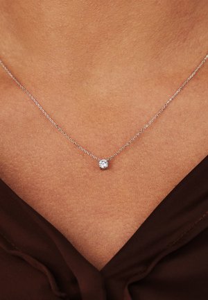 Collier en chaîne en argent avec un petit pendentif rond en pierre transparente, reposant sur un décolleté de teinte chair avec un haut marron visible.