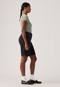 Γυναίκα με πλεξούδες που φοράει πράσινο ριγωτό μπλουζάκι, μαύρες τζιν βερμούδες μέχρι το γόνατο, άσπρες κάλτσες και μαύρα μοκασίνια, στέκεται πλάγια.