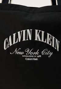 Μαύρη τσάντα tote από καμβά με λευκό κεντημένο κείμενο: "CALVIN KLEIN New York City established in 1968," με απλό σχέδιο και ιμάντες.