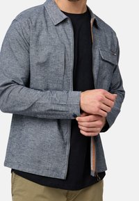 Homme ajustant le poignet d'une veste-chemise grise dézippée par-dessus un t-shirt noir, portant un pantalon kaki, sur un fond uni.