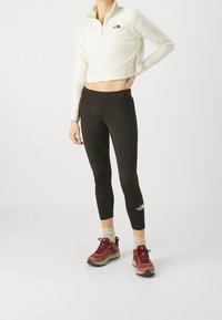 Felpa leggera a zip cropped color crema, leggings neri e scarpe da ginnastica marrone-rossicce con suola testurizzata. I leggings presentano un dettaglio con logo sul lato.
