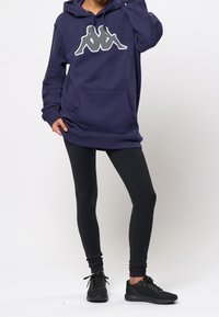Personne portant un sweat à capuche bleu marine avec un logo blanc, des leggings noirs et des baskets noires, debout avec une main sur la hanche et l'autre touchant la tête.