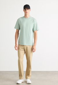 T-shirt de algodão verde menta com mangas curtas, combinada com calças bege e ténis brancos. O conjunto é simples, sem gráficos visíveis.
