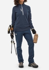 Kobieta w granatowej polarowej bluzie i spodniach, trzymająca kijki trekkingowe w jednej ręce i butelkę wody w drugiej, gotowa do wędrówki.