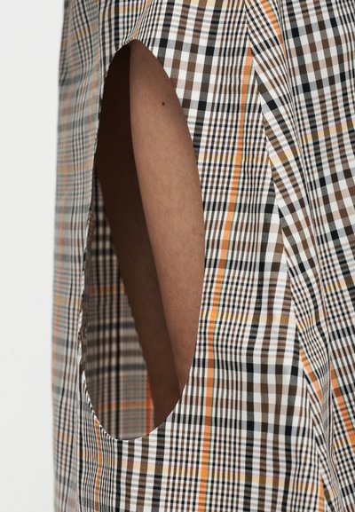 Καφέ δέρμα ορατό μέσα από οβάλ κοπή σε καρό ύφασμα με καφέ, μαύρες, λευκές και πορτοκαλί διασταυρούμενες γραμμές.