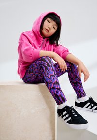 Rosa Kapuzenpullover mit durchgehendem Reißverschluss vorne, kombiniert mit schwarzen Leggings mit violetten und pinkfarbenen abstrakten Mustern sowie schwarzen Turnschuhen.