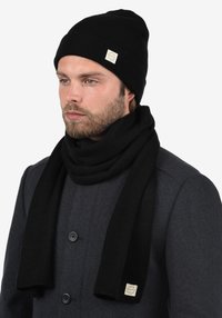 Zwart geribbelde muts en sjaal set gemaakt van gebreide stof. Voorzien van een vierkante label op de sjaal. Gedragen met een donkergrijze jas.