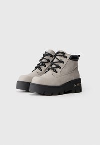 Botas de tobillo de ante beige con suelas de goma gruesa negras, cuello acolchado negro y cierre de cordones negro sobre un fondo blanco.