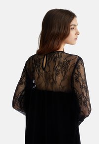 Abito nero con corpetto in pizzo decorato con motivi floreali e maniche lunghe trasparenti. Il gonnellino è realizzato in morbido velluto, aggiungendo texture.