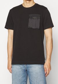 Svart bomullströja med rund hals, korta ärmar och en framficka. Fickan har ett lock och en diskret logotypdetalj.