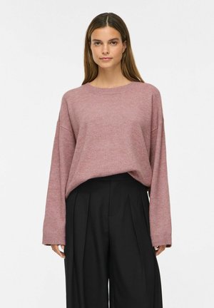 Kvinde med glat brunt hår iført en løs mauve sweater og brede sorte bukser, stående mod en ensfarvet hvid baggrund.