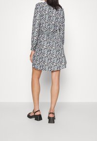 Vestido envolvente floral de manga larga, con un fondo oscuro, patrón de flores multicolor y una cintura ceñida. Sandalias negras de tacón cuadrado.