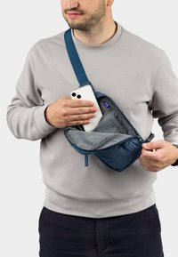 Borsa a tracolla realizzata in tessuto resistente di colore blu scuro, dotata di una tasca con zip. L'utente tiene uno smartphone mentre indossa un maglione grigio.