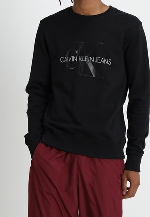 Mann trägt schwarzen Calvin Klein Jeans Sweatshirt und lockere rote Hose, steht mit einer Hand in der Tasche.