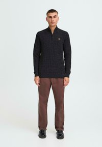 Dunkelgrauer, strukturierter Strickpullover mit einem Reißverschlusskragen, kombiniert mit braunen Hosen und schwarzen Schuhen. Mann steht vor einem neutralen Hintergrund.