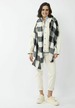 Witte denimjack over een lichtblauw shirt, witte broek en beige platform sneakers. Gewikkeld in een zwart-wit geruite sjaal.