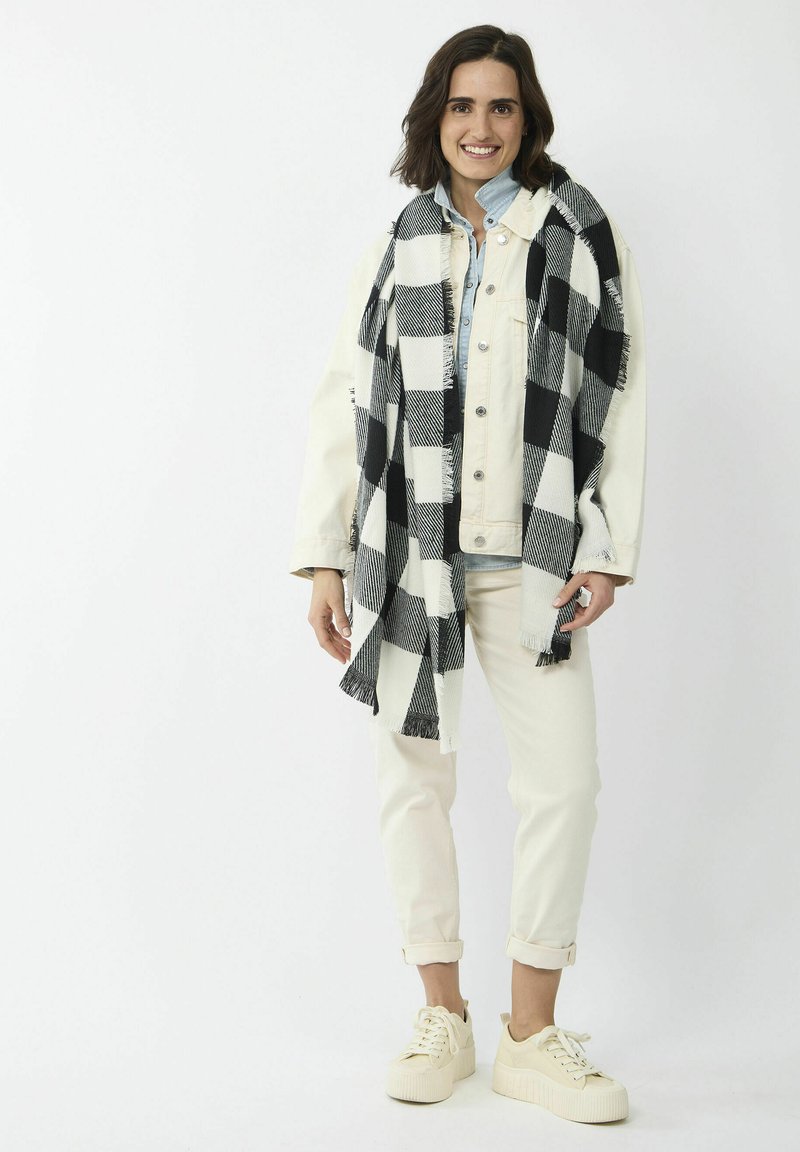 Witte denimjack over een lichtblauw shirt, witte broek en beige platform sneakers. Gewikkeld in een zwart-wit geruite sjaal.